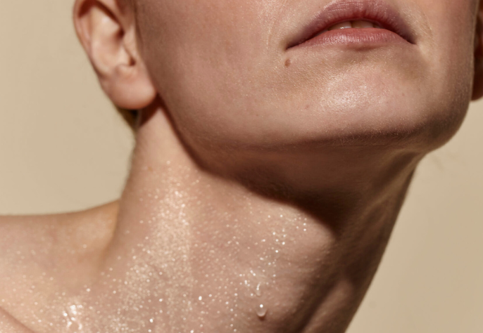 A close-up of a person's neck, jawline, and chin, showing glistening skin with a water droplet, conveying deep hydration and a healthy glow.
