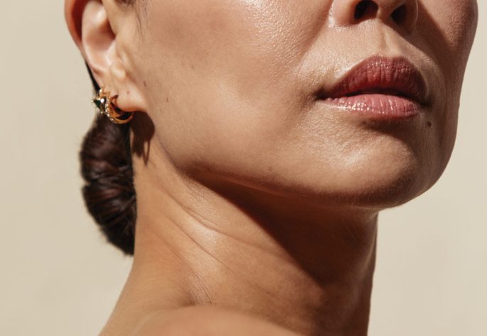 A close-up profile of a person's jawline, neck, and lower face, showcasing smooth, healthy skin with a subtle glow, and a small earring in their ear.