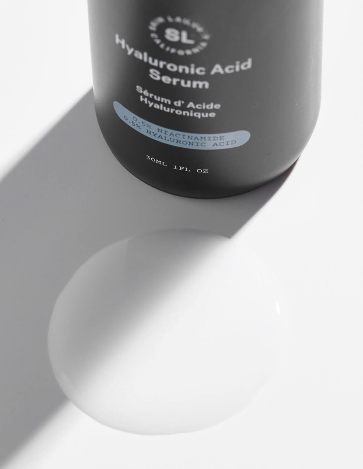A close-up of a black dropper bottle of Skin Laundry Hyaluronic Acid Serum, with its label showing "0.5% NIACINAMIDE 0.5% HYALURONIC ACID," casting a long shadow onto a white surface with a circular light reflection.