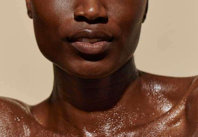 A close-up of a person's neck, jawline, and bare shoulders with deep brown, glistening skin and visible water droplets, emphasizing intense hydration and a healthy glow.