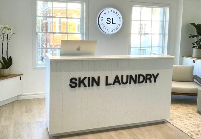 The bright and pristine white reception desk of a Skin Laundry clinic, featuring the "SKIN LAUNDRY" name on the front, an Apple laptop, and a circular illuminated "SKIN LAUNDRY CALIFORNIA SL" sign mounted on the wall above, flanked by large windows.
