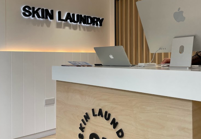 A close-up of the Skin Laundry reception desk, featuring a MacBook laptop, an Apple iMac desktop, and the "SKIN LAUNDRY" logo prominently displayed on both the wooden desk and the illuminated wall signage.