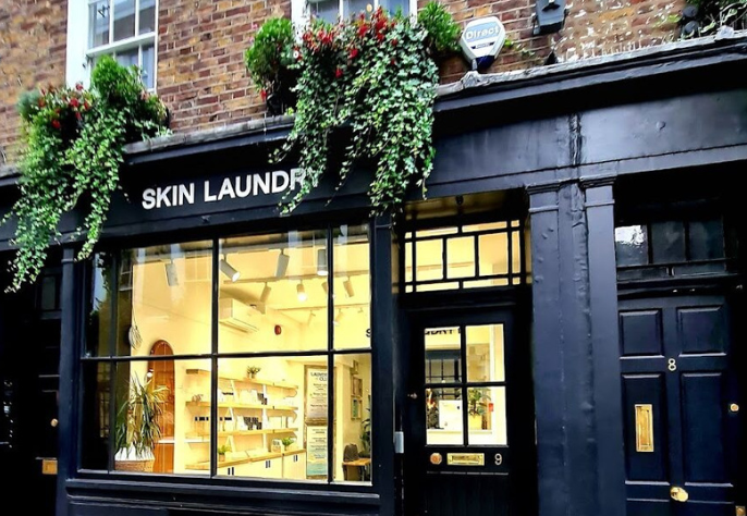 The exterior of a Skin Laundry clinic storefront in a classic brick building, featuring a black facade with large display windows, the "SKIN LAUNDRY" name, and decorative hanging plants.