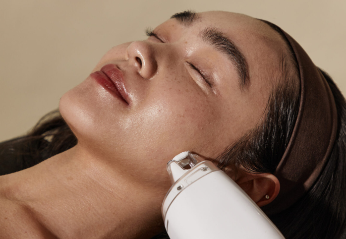 A person with a brown headband and closed eyes, looking relaxed while receiving a facial treatment with a white handheld device being applied to their cheek, emphasizing a gentle skincare procedure.