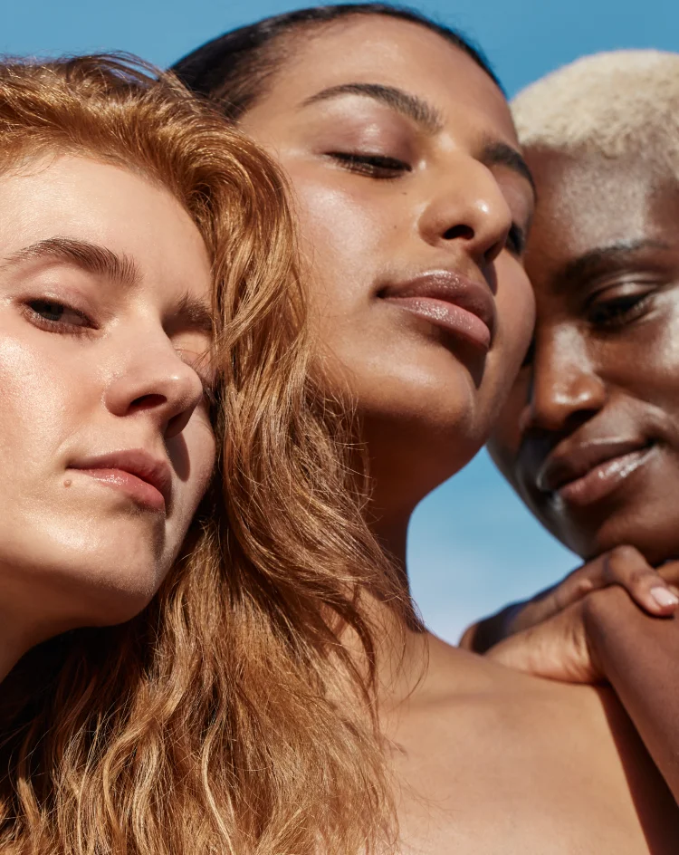 A close-up group portrait of three diverse individuals with healthy, glowing skin, featuring varying complexions and hair colors, looking serene and connected against a clear blue sky.