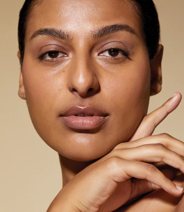 A close-up portrait of a person with warm, healthy-looking skin, looking directly at the camera with one hand gently touching their chin, highlighting their natural complexion and clear skin.