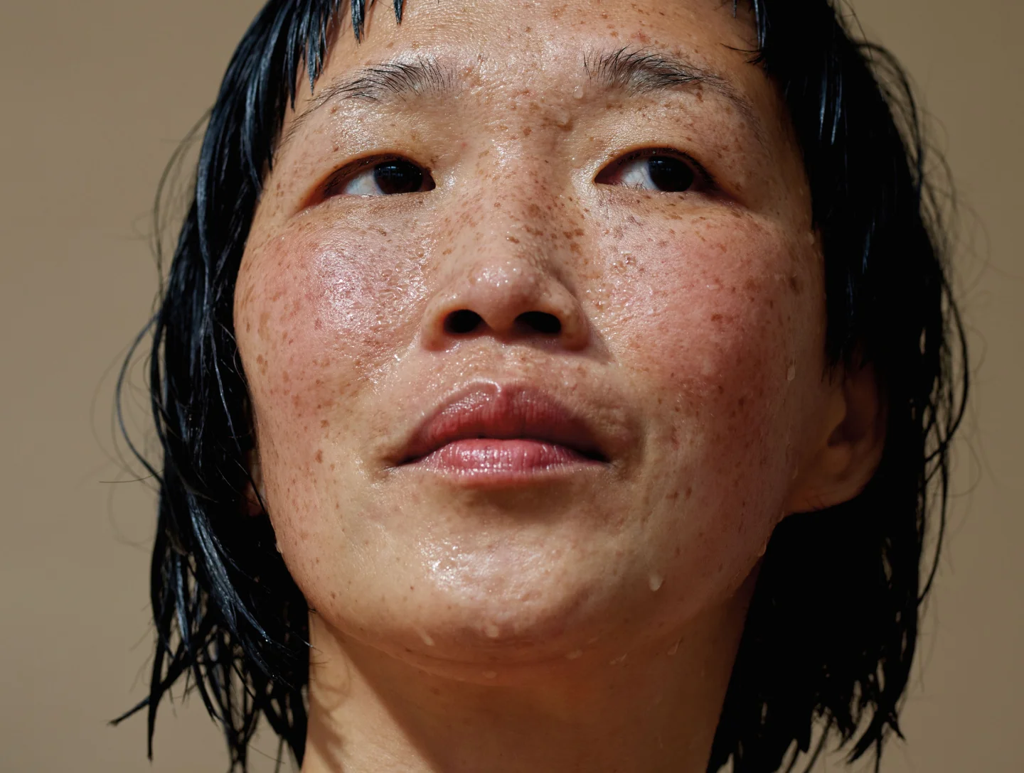 A close-up portrait of a person with short dark hair and freckles, with wet, glistening skin and water droplets on their face, looking upwards.