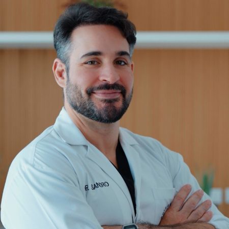 A smiling male professional with a beard, wearing a white lab coat with "DR. SANDRO" partially visible, standing with his arms crossed and looking directly at the camera.
